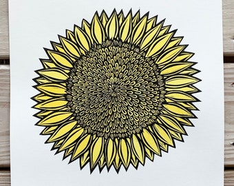 May include: A black and yellow linocut print of a sunflower. The print is titled "Sunflower" and is dated 2019. The artist's signature is in the bottom right corner.