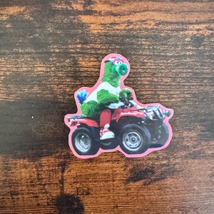 May include: A sticker featuring a green character wearing a white shirt, red pants, and a helmet, riding a red ATV. The sticker has a pink border and is set against a brown wooden surface.