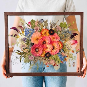 May include: A rectangular dark wood floating frame containing a pressed flower arrangement. The display features a vibrant mix of dried pink, orange, and blue blossoms, along with green foliage and stems, preserved between two clear glass panes.