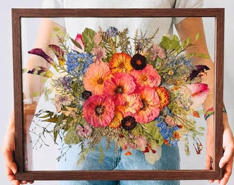 May include: A rectangular dark wood floating frame containing a pressed flower arrangement. The display features a vibrant mix of dried pink, orange, and blue blossoms, along with green foliage and stems, preserved between two clear glass panes.