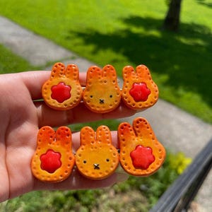 May include: Six orange and red bunny-shaped polymer clay pins. Each pin has a round body with a red flower-shaped center. The bunnies have small black eyes and a stitched mouth. The pins are handmade.