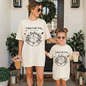 May include: Two people wearing matching cream-colored t-shirts with the text "Happy Earth Day" and a graphic of the Earth held in hands. The shirts have a relaxed fit. One person holds a drink with a straw.