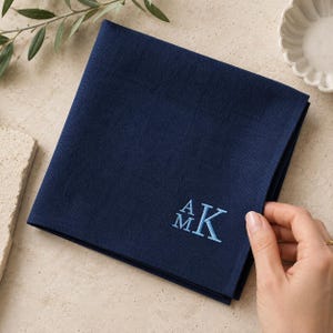 May include: A square navy blue fabric napkin featuring a light blue embroidered monogram with the letters A, M, and K. The fabric has a visible woven texture and is folded neatly. The embroidery is positioned in the lower right corner.