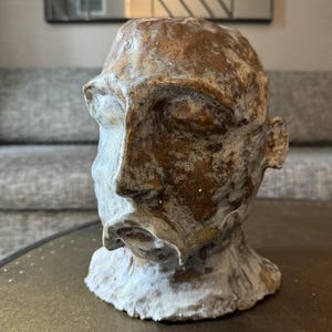 May include: A ceramic sculpture of a human head with a textured, mottled finish in shades of brown, bronze, and off-white. The face features closed eyes, a prominent nose, and a defined mouth, with a rough, uneven surface texture throughout.