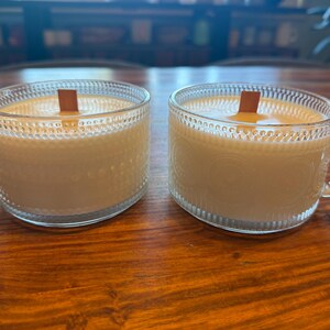以下が含まれることがあります： 木製の芯と淡いベージュ色のワックスが入った、ガラス製のティーカップキャンドルが2つ。透明なガラスカップにはテクスチャパターンとハンドルが付いています。キャンドルは濃い木製の表面に置かれています。