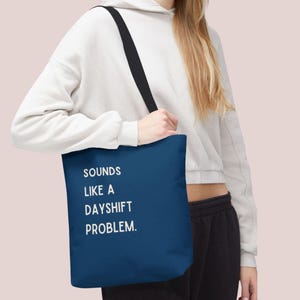 Könnte beinhalten: Marineblaue Tragetasche mit dem weißen Text "SOUNDS LIKE A DAYSHIFT PROBLEM". Die Tasche hat einen schwarzen Riemen und wird getragen. Ein einfaches, alltägliches Accessoire.