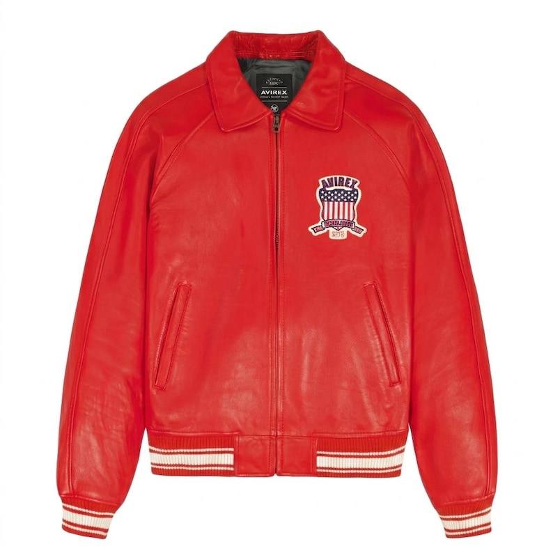 Puede incluir: Chaqueta de cuero rojo brillante con cuello y cierre de cremallera. La chaqueta presenta un parche en el pecho con el texto "AVIREX" y un dise&ntilde;o que incorpora la bandera estadounidense. Los pu&ntilde;os y el dobladillo tienen detalles a rayas blancas y rojas.