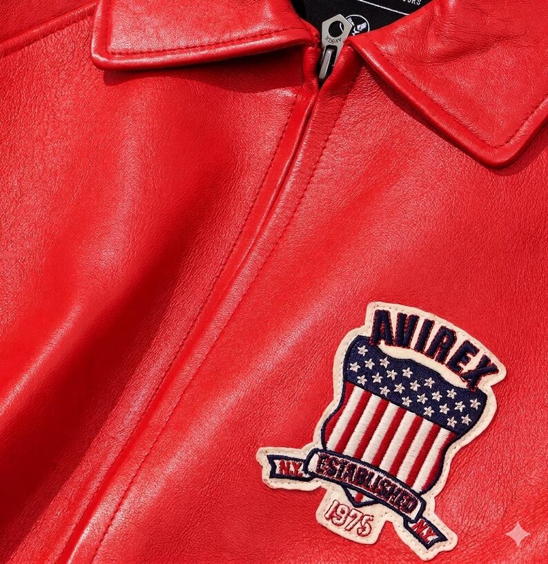 Puede incluir: Primer plano de una chaqueta de cuero roja con cuello y cremallera. La chaqueta presenta un parche con la palabra "AVIREX" sobre un dise&ntilde;o de la bandera estadounidense y el texto "NY ESTABLISHED 1975".