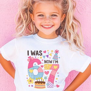 May include: A white t-shirt with the text "I WAS 67 NOW I'M" and a colorful graphic. The graphic includes the number 67, a birthday cake, and festive decorations. The shirt is worn by a young girl with a pink bow in her hair.