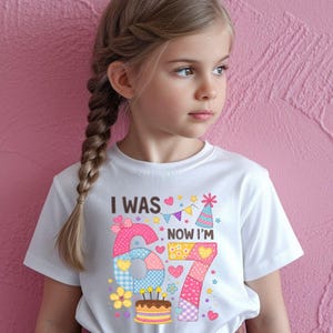 May include: A white t-shirt with the text "I WAS 6 NOW I'M 7" in colorful patchwork style. The design includes a birthday cake, hearts, and stars. The shirt is worn by a young girl with a braided hairstyle.