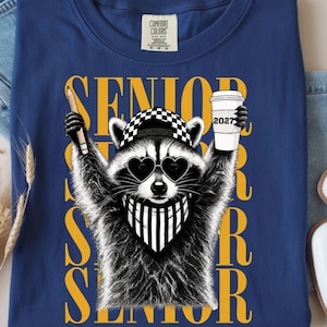 Pu&ograve; includere: Una t-shirt blu con la grafica di un procione che indossa un berretto a scacchi, occhiali da sole a forma di cuore e una bandana a righe. Il procione tiene in mano una matita e un bicchiere con la scritta 2027. La parola SENIOR &egrave; stampata in grandi lettere gialle.