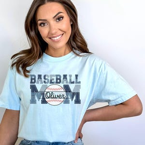May include: Light blue t-shirt with the words "BASEBALL MOM" in a layered blue design. A baseball graphic with the name "Oliver" is centered below the word "MOM".