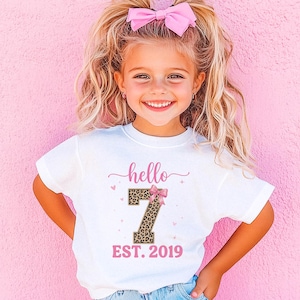 May include: A young girl wearing a white t-shirt with the text "hello 7 EST. 2019" in pink. The number 7 has a leopard print design and a pink bow. The girl has blonde hair and is wearing a pink bow.