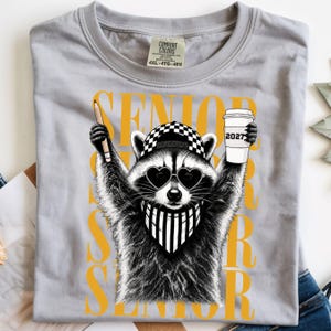 Pu&ograve; includere: Una t-shirt grigio chiaro con la grafica di un procione che indossa un berretto da baseball a scacchi, occhiali da sole a forma di cuore e una bandana a righe. Il procione tiene in mano una matita e un bicchiere di caff&egrave; con scritto 2027. SENIOR &egrave; in giallo.