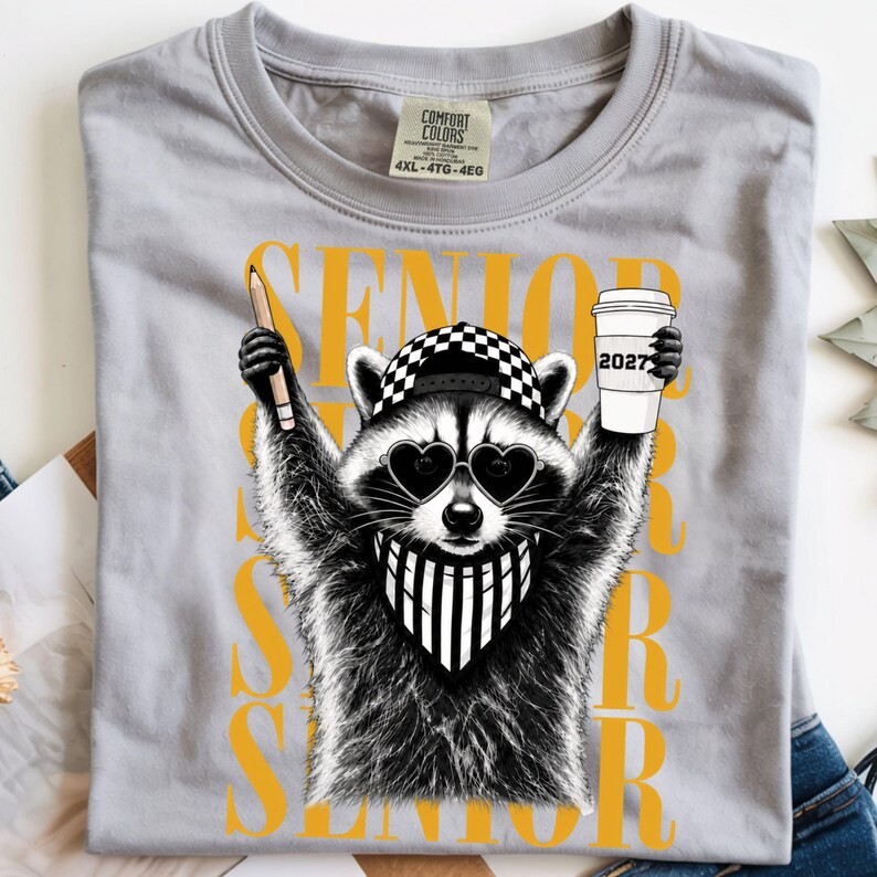 Pu&ograve; includere: Una t-shirt grigio chiaro con la grafica di un procione che indossa un berretto da baseball a scacchi, occhiali da sole a forma di cuore e una bandana a righe. Il procione tiene in mano una matita e un bicchiere di caff&egrave; con scritto 2027. SENIOR &egrave; in giallo.