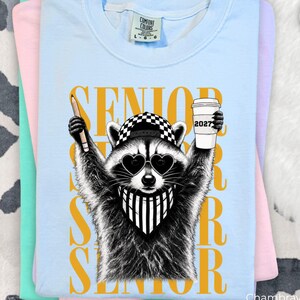 Pu&ograve; includere: Una t-shirt azzurra con la grafica di un procione che indossa un berretto da baseball a scacchi, occhiali da sole a forma di cuore e una bandana a righe. Il procione tiene in mano una matita e un bicchiere con la scritta 2027. La parola SENIOR &egrave; dorata.