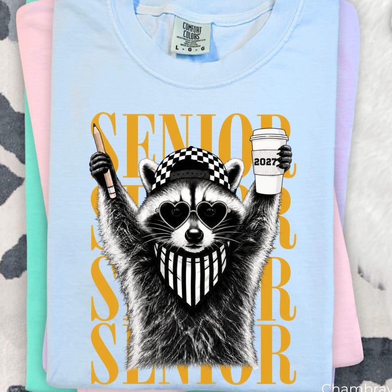 Pu&ograve; includere: Una t-shirt azzurra con la grafica di un procione che indossa un berretto da baseball a scacchi, occhiali da sole a forma di cuore e una bandana a righe. Il procione tiene in mano una matita e un bicchiere con la scritta 2027. La parola SENIOR &egrave; dorata.