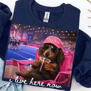 May include: A navy blue sweatshirt featuring a graphic of a dachshund wearing sunglasses and a pink baseball cap that says Ellie's Mom, sitting in a pink folding chair holding a plastic cup with a straw, with the text I live here now below.