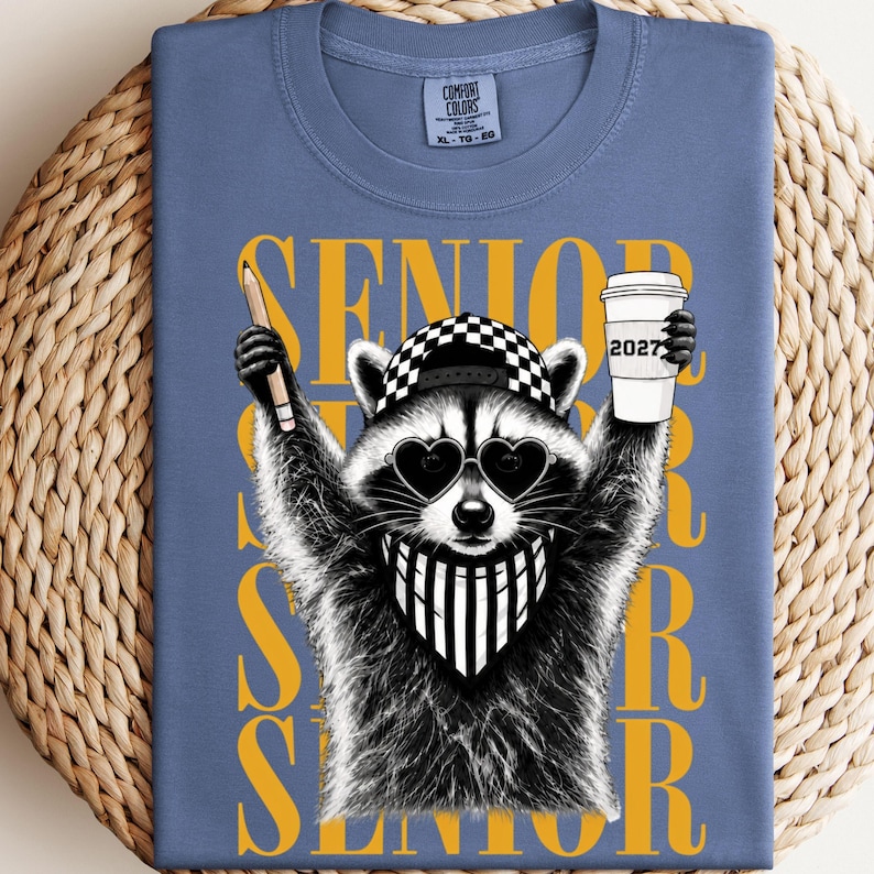 Pu&ograve; includere: Una t-shirt blu con la grafica di un procione che indossa un berretto a scacchi, occhiali da sole a forma di cuore e una bandana a righe. Il procione tiene in mano una matita e un bicchiere con la scritta 2027. La parola SENIOR &egrave; stampata in giallo.