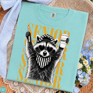 Pu&ograve; includere: Una t-shirt azzurra con la grafica di un procione che indossa occhiali da sole a forma di cuore, un berretto a scacchi e una bandana a righe. Il procione tiene in mano una matita e un bicchiere con la scritta 2027. La parola SENIOR &egrave; in giallo.