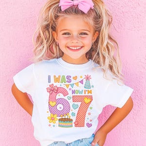 May include: White t-shirt with the text "I WAS 6 NOW I'M 7" in colorful, patchwork-style letters and graphics. The design includes a birthday cake, hearts, and a party hat. The shirt is worn by a young girl with blonde hair and a pink bow.