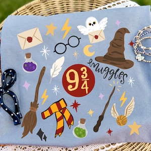 May include: A light blue t-shirt featuring a graphic print of magical items including a brown sorting hat, a broomstick, a wand, a golden snitch, round glasses, a red and yellow scarf, potion bottles, an owl, and the text 9 3/4 and muggles.
