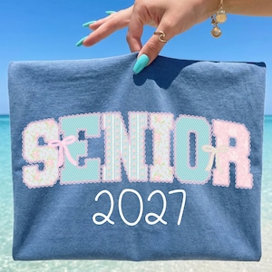 May include: A blue t-shirt featuring the word SENIOR in large, patterned block letters with floral, polka dot, and striped designs in shades of pink and light blue, accented with small bows. Below the word, the number 2027 is printed in white.