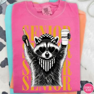 Pu&ograve; includere: Una t-shirt rosa acceso con la grafica di un procione che indossa un berretto da baseball a scacchi, occhiali da sole a forma di cuore e una bandana a righe. Il procione tiene in mano una matita e un bicchiere di caff&egrave; con scritto 2027.