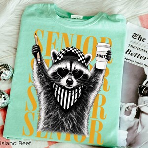 Pu&ograve; includere: Una t-shirt verde chiaro con la grafica di un procione che indossa occhiali da sole a forma di cuore, un berretto a scacchi e una bandana a righe. Il procione tiene in mano una matita e un bicchiere con la scritta 2027. Testo giallo: SENIOR.