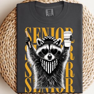 Pu&ograve; includere: Una t-shirt grigio antracite con la grafica di un procione che indossa occhiali da sole a forma di cuore, un berretto a scacchi e una bandana a righe. Il procione tiene in mano una matita e un bicchiere con la scritta 2027. La parola SENIOR &egrave; gialla.