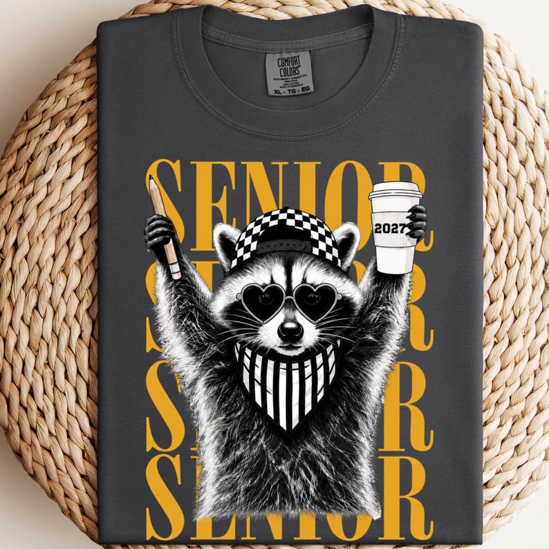 Pu&ograve; includere: Una t-shirt grigio antracite con la grafica di un procione che indossa occhiali da sole a forma di cuore, un berretto a scacchi e una bandana a righe. Il procione tiene in mano una matita e un bicchiere con la scritta 2027. La parola SENIOR &egrave; gialla.