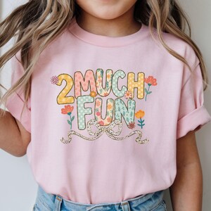 May include: A light pink t-shirt with the text "2 MUCH FUN" in colorful, floral-patterned letters. The number "2" is yellow, and the word "FUN" is in a blue floral pattern. A dotted ribbon is at the bottom.