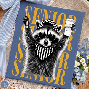 Pu&ograve; includere: Una stampa rettangolare blu con un procione che indossa un berretto a scacchi, occhiali da sole a forma di cuore e una bandana a righe. Il procione tiene in mano una matita e un bicchiere con la scritta 2027. Sullo sfondo appare la parola SENIOR in giallo.