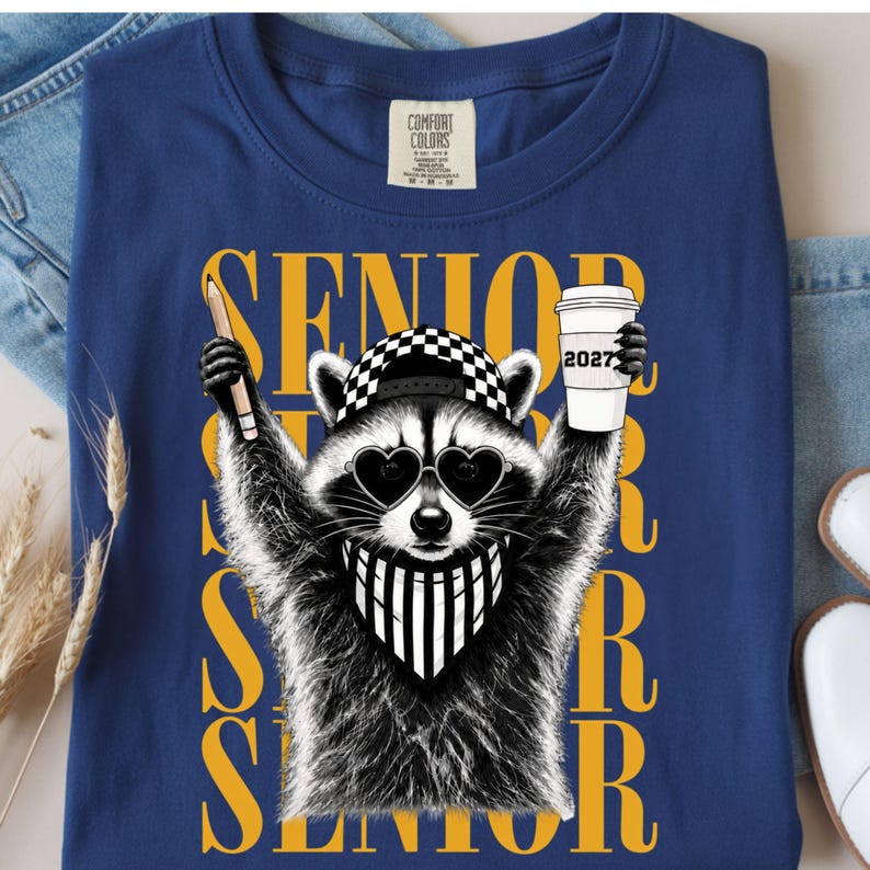 Pu&ograve; includere: Una t-shirt blu con la grafica di un procione che indossa un berretto a scacchi, occhiali da sole a forma di cuore e una bandana a righe. Il procione tiene in mano una matita e un bicchiere con la scritta 2027. La parola SENIOR &egrave; stampata in grandi lettere gialle.