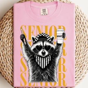Pu&ograve; includere: Una t-shirt rosa chiaro con la grafica di un procione che indossa un berretto da baseball a scacchi bianchi e neri, occhiali da sole a forma di cuore e una bandana a righe. Il procione tiene in mano una matita e un bicchiere di caff&egrave; con la scritta 2027. La parola SENIOR &egrave; stampata in grandi lettere gialle sullo sfondo.