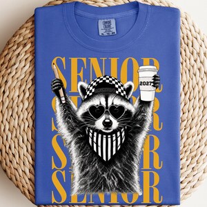 Pu&ograve; includere: Una t-shirt blu con la grafica di un procione che indossa un berretto a scacchi, occhiali da sole a forma di cuore e una bandana a righe. Il procione tiene in mano una matita e un bicchiere con la scritta 2027. Il testo SENIOR &egrave; in giallo.