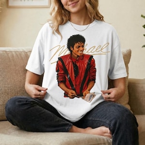 May include: White t-shirt featuring a graphic of Michael Jackson in a red jacket with black accents. The shirt has the name "Michael" in gold script across the top. The image is a portrait of the singer.