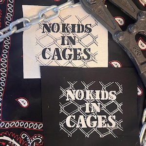 May include: Two patches with the text "NO KIDS IN CAGES" in white, one on a black background and one on a cream background. A silver chain and a bolt cutter are also in the image. The background is a black bandana with a paisley pattern.