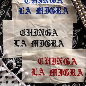 May include: Three off-white fabric patches with the text "CHINGA LA MIGRA" in blue, black, and red gothic lettering. The patches are on a black and white paisley background with a studded belt and a fringed scarf.