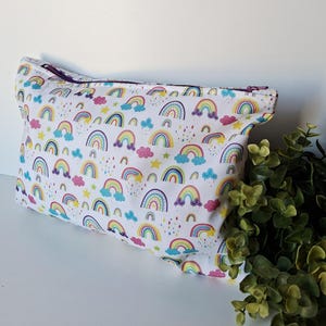 Puede incluir: Un neceser blanco con un estampado repetido de arcoíris, estrellas y nubes de colores. La bolsa tiene una cremallera morada y está hecha de tela. La bolsa es rectangular y tiene una parte superior redondeada.