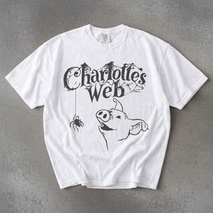 May include: White t-shirt featuring a black graphic of a pig looking up at a spider hanging from the text "Charlotte's Web." The text is arched above the pig, with a spider web design. The t-shirt has short sleeves and a crew neck.