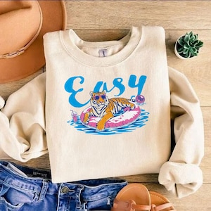 May include: Cream-colored sweatshirt with the word "Easy" in blue script. A cartoon tiger wearing sunglasses floats on a pink inner tube in water. A drink and a beach ball are also in the design.