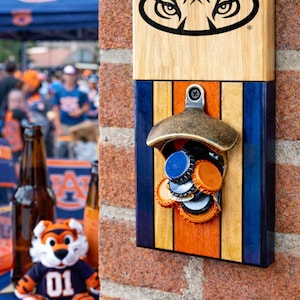 May include: Wooden wall-mounted bottle opener featuring a black and white tiger eye logo. The opener has a brass finish and is mounted on a wood panel with blue, orange, and natural wood stripes. Several bottle caps are held by the opener.