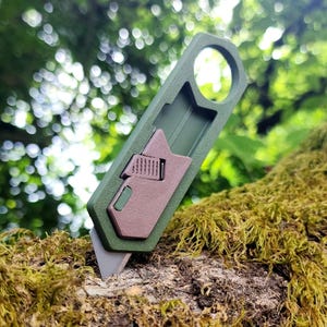 May include: A utility knife with an olive green frame and a brown sliding blade mechanism. The tool features a circular hole at the top of the handle and a sharp, metallic blade tip extending from the bottom, designed for precision cutting tasks.