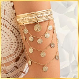 May include: Gold-toned arm cuff with a wide band featuring pyramid studs. Delicate chains cascade down, adorned with coin-shaped charms. The jewelry is worn on the upper arm, showcasing a bohemian style.