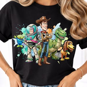 May include: A black short-sleeved t-shirt featuring a colorful graphic print of five animated characters including a space ranger, a cowboy, a green dinosaur, a dachshund, and a small green alien, set against a vibrant watercolor splash background.