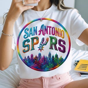 May include: A white t-shirt featuring a circular graphic with a rainbow border. The text reads San Antonio Spurs in a stylized font, with the team logo replacing the letter U. Below the text is a colorful, detailed cityscape silhouette.