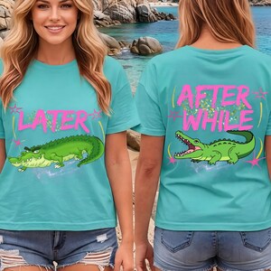 Könnte beinhalten: Türkisfarbene T-Shirts mit einem Cartoon-Alligator-Motiv. Auf der Vorderseite des Shirts steht "LATER" in Pink, und auf der Rückseite steht "AFTER WHILE" in Pink. Der Alligator ist grün mit gelben Akzenten.