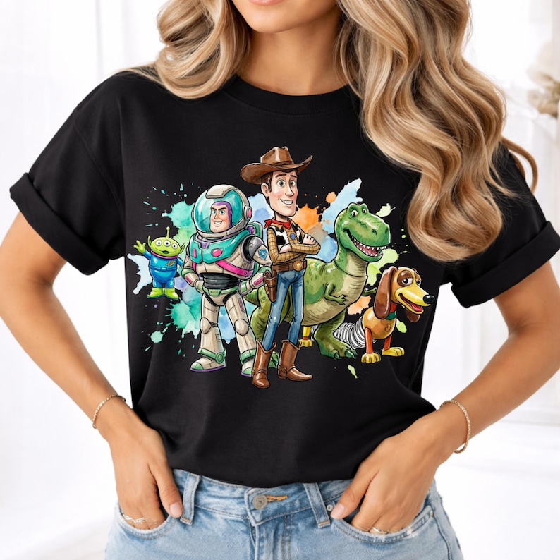 May include: A black short-sleeved t-shirt featuring a colorful graphic print of five animated characters including a space ranger, a cowboy, a green dinosaur, a dachshund, and a small green alien, set against a vibrant watercolor splash background.