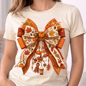 Pode incluir: Uma t-shirt de cor creme com um grande gráfico central de um laço decorativo. O laço apresenta um padrão de abóboras cor de laranja, folhas de outono, bolotas e bagas. Pendurados no laço estão berloques que incluem um cachecol, um cogumelo e um copo com a etiqueta PUMPKIN SPICE.